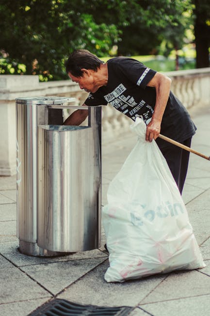 A middle-aged man with dark hair is seen disposing of waste into a metallic, cylindrical waste bin located on a paved sidewalk. He is bent slightly forward, wearing a dark t-shirt with white and blue 